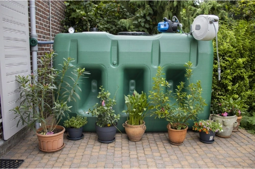 Bovengrondse kunststof regenwatertank van 3000 liter met pomp en oprolslanghaspel, aangesloten op de regenpijp en omringd door bloempotten in een tuin.