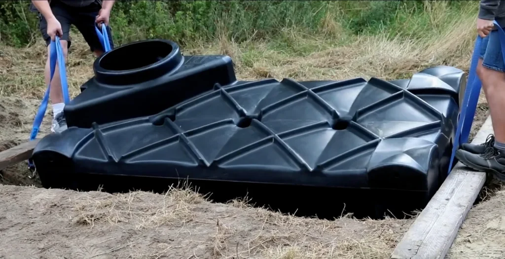 Stappenplan plaatsen van regenwaterput. Twee personen laten een lichte kunststof regenwaterput zakken in een gegraven put met behulp van spanbanden.